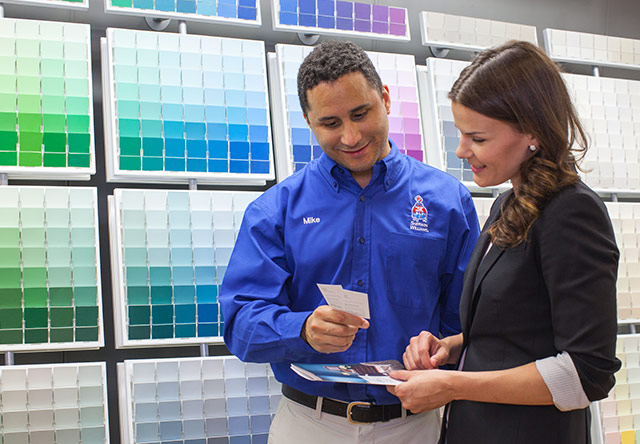 A Sherwin-Williams employee holding two color chips while conversing with a woman in a dark blazer in front of a color chip wall.